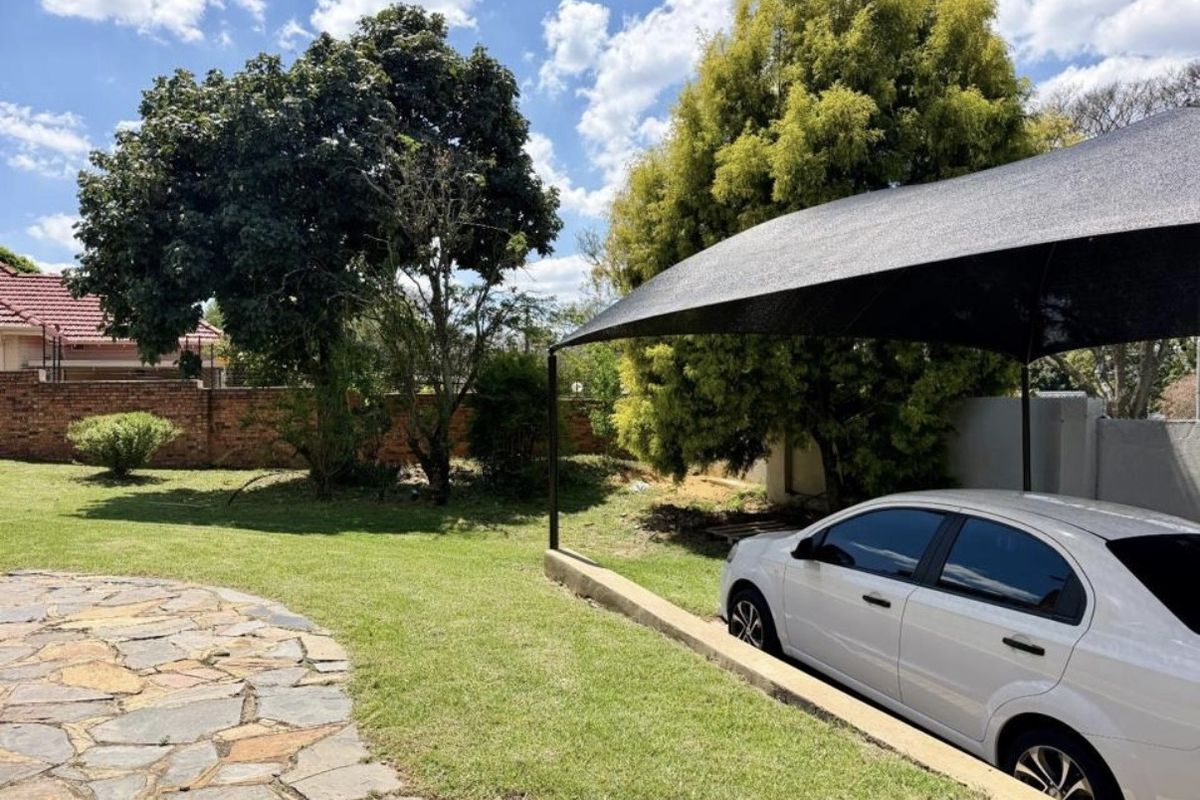 A rear garden area featuring a stone paved path, a lawn, and a covered parking space with a shade sail structure.