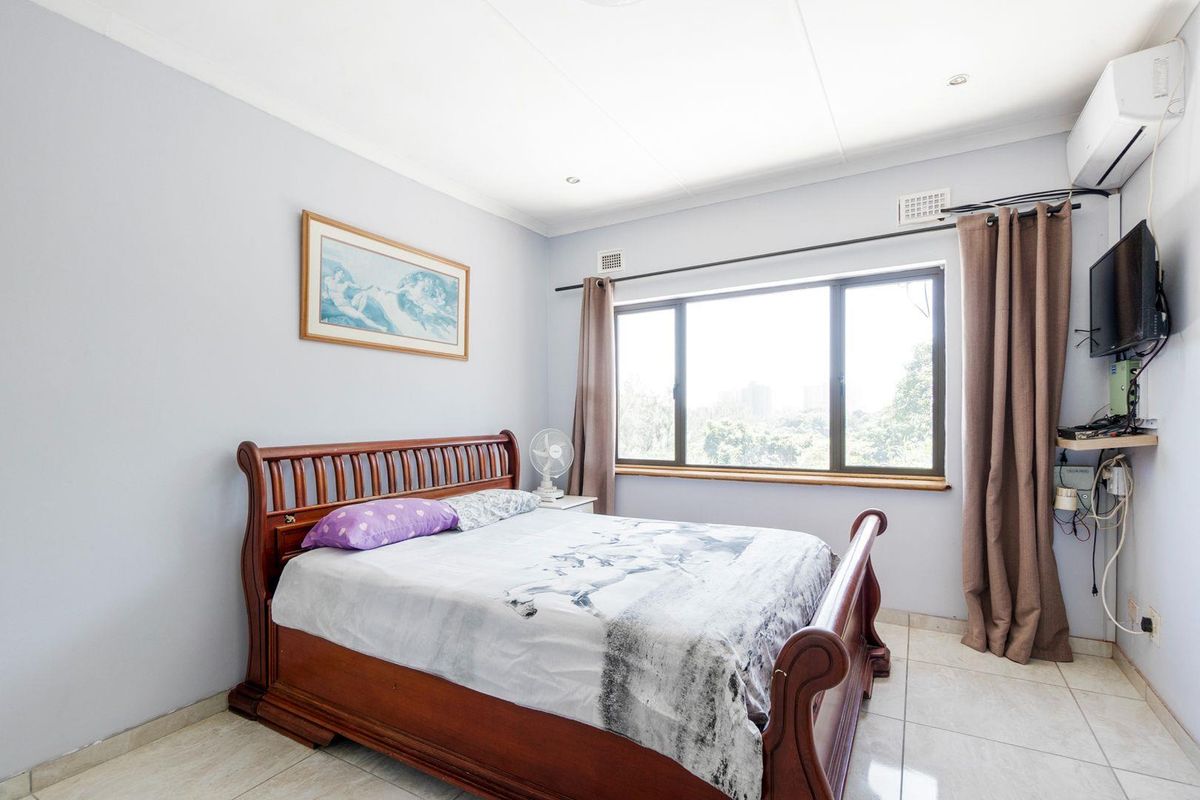 A well-lit bedroom featuring tiled flooring, a large window with natural light, and a wall-mounted air conditioning unit.