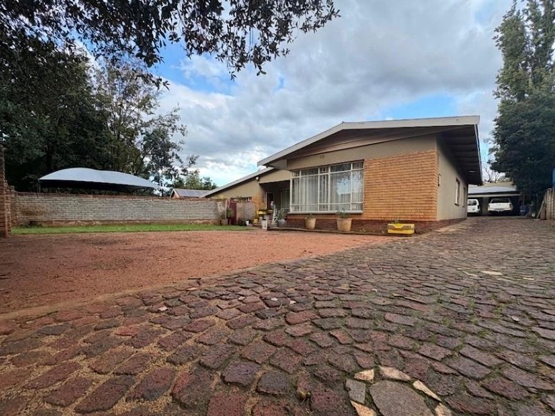 Front exterior view of the house featuring a large cobblestone driveway, brick boundary wall, and a carport, set on a spacious plot.