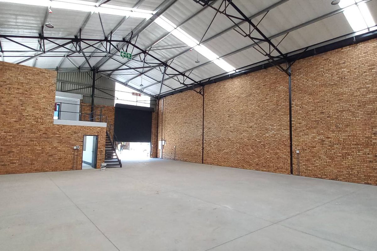 Spacious industrial warehouse featuring high ceilings with exposed steel trusses, concrete flooring, brick walls, a mezzanine level, and a large roller shutter door.