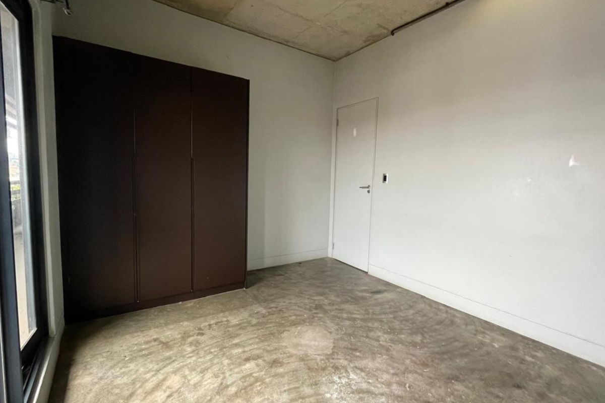 A minimalist bedroom featuring exposed concrete ceilings, polished concrete flooring, and a built-in dark wood wardrobe with access to an adjacent balcony.