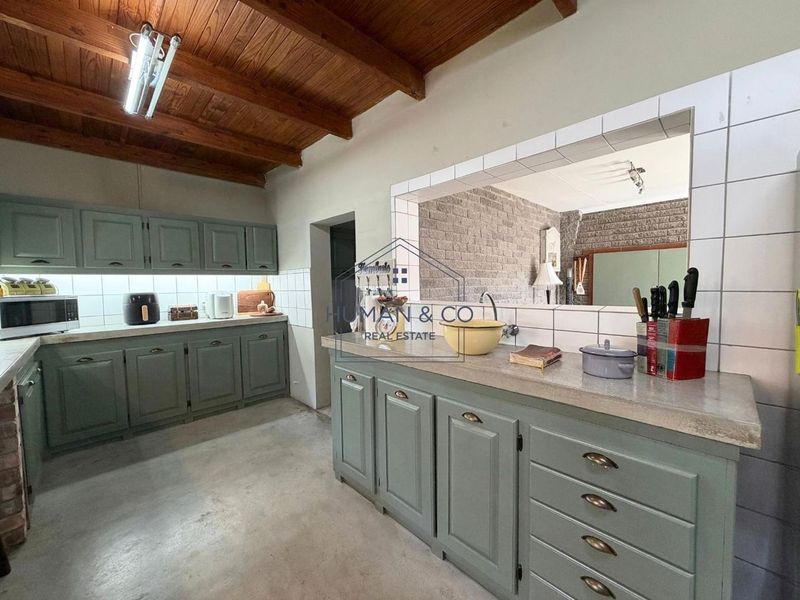A functional kitchen featuring sage green cabinetry, stone countertops, a tiled backsplash, and an exposed timber beam ceiling with a convenient pass-through window.