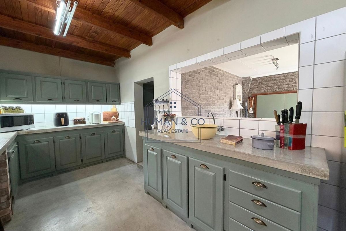 A functional kitchen featuring sage green cabinetry, stone countertops, a tiled backsplash, and an exposed timber beam ceiling with a convenient pass-through window.