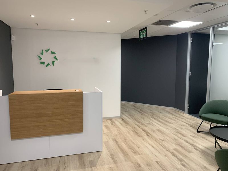 Modern office reception area featuring a clean reception desk, laminate wood-look flooring, recessed lighting, and a glass-partitioned office entrance.