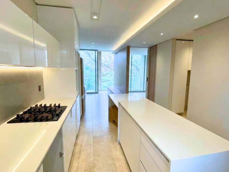 Modern, minimalist kitchen featuring sleek white cabinetry, stone countertops, integrated gas cooktop, and recessed lighting, leading into an open-plan living area.