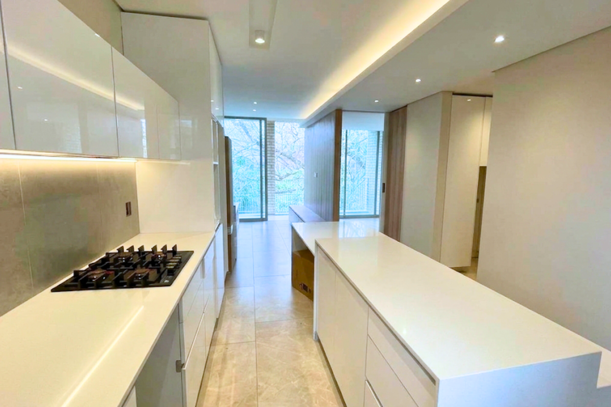 Modern, minimalist kitchen featuring sleek white cabinetry, stone countertops, integrated gas cooktop, and recessed lighting, leading into an open-plan living area.