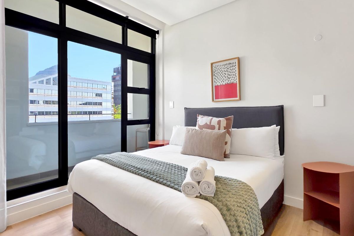 Modern bedroom featuring light laminate flooring and large floor-to-ceiling windows that provide ample natural light and access to an outdoor balcony area.