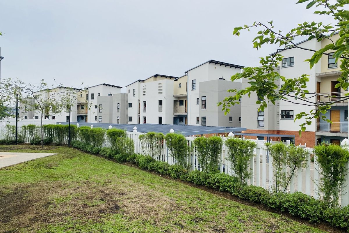 A view of a modern multi-story apartment complex featuring a well-maintained garden, white picket fencing, and covered carport parking facilities.