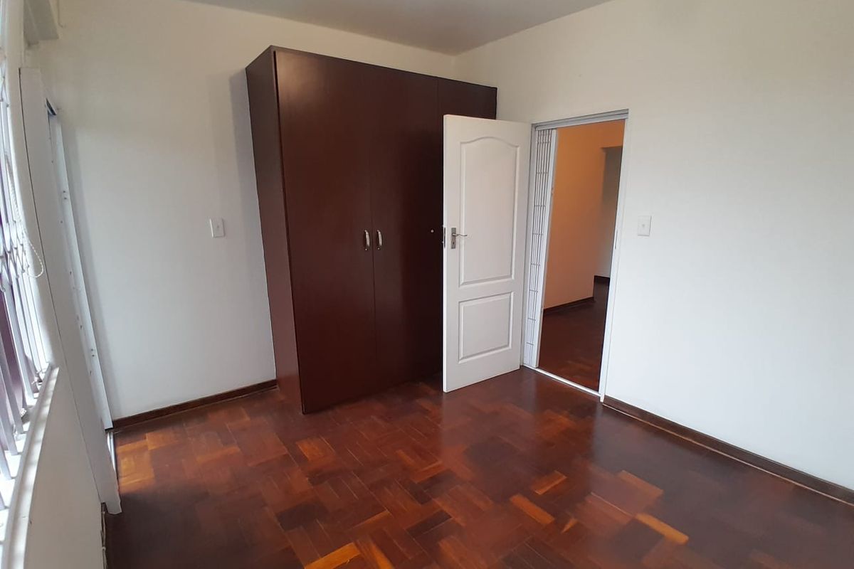 A bright, empty bedroom featuring classic parquet flooring and a built-in dark wood wardrobe, offering a clean canvas for personalization.