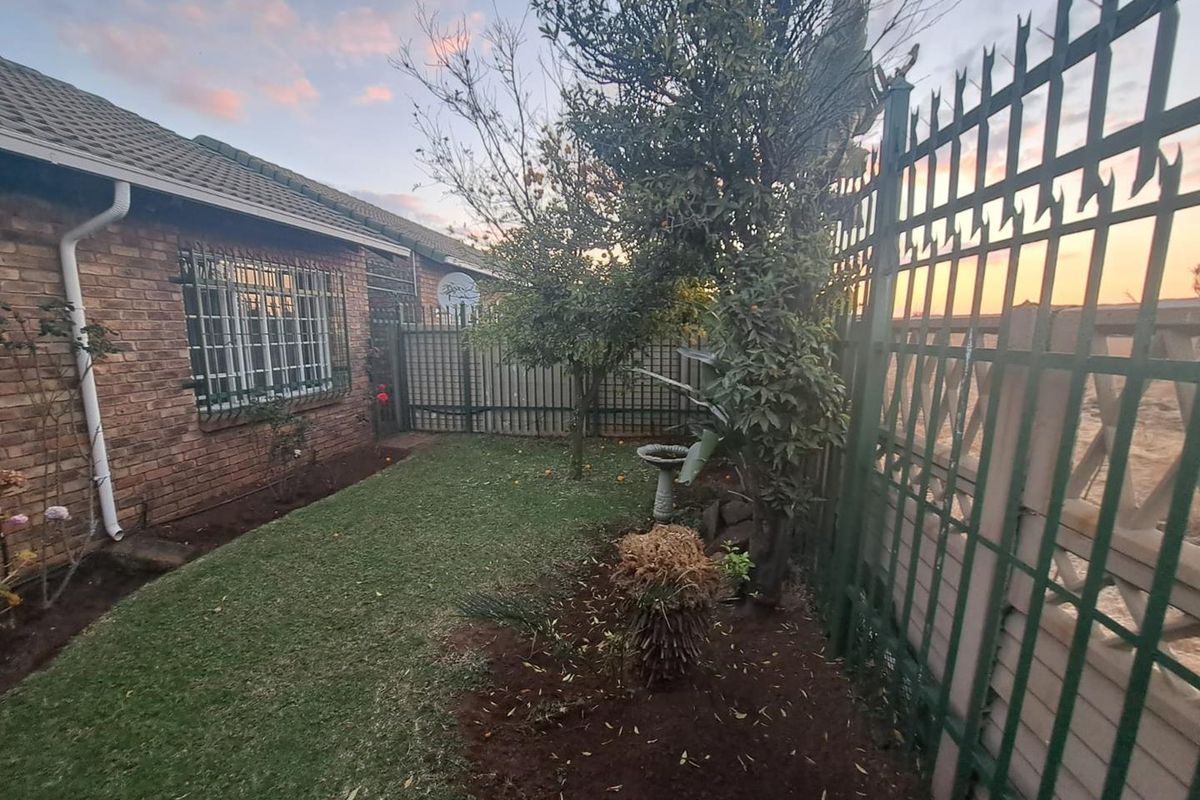 A rear garden area featuring a brick townhouse exterior, perimeter metal fencing, and established trees, providing a private outdoor space.