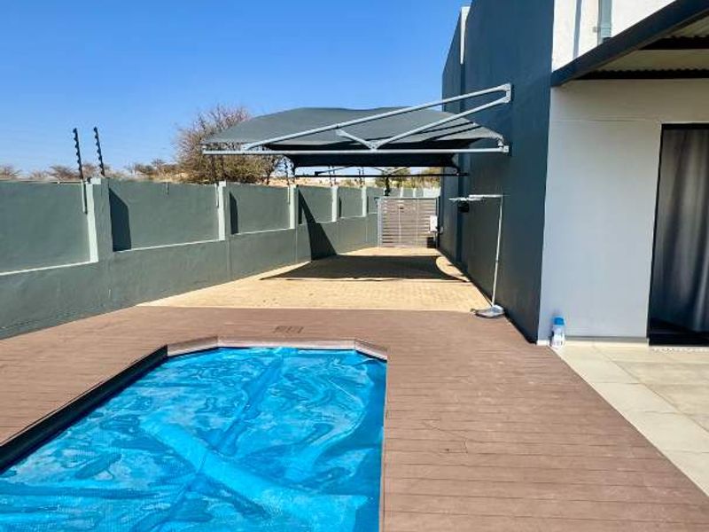 Outdoor pool area featuring timber decking, a swimming pool with cover, paved driveway, and a shade sail structure against a modern townhouse exterior.