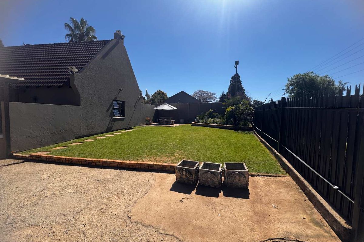 A private rear garden featuring a well-maintained lawn, brick-edged garden beds, and secure perimeter fencing, adjacent to a concrete driveway area.