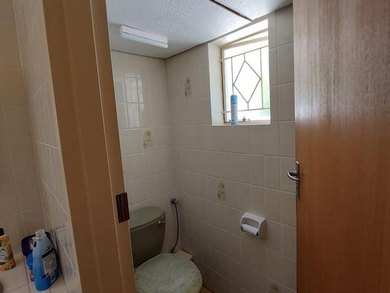 A compact, dated bathroom featuring ceramic wall and floor tiles, a standard toilet, and a small window with a security grille.
