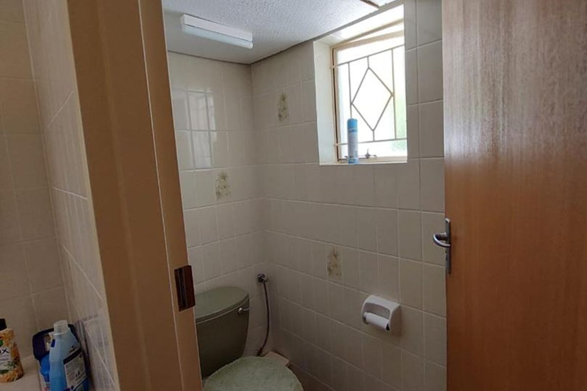 A compact, dated bathroom featuring ceramic wall and floor tiles, a standard toilet, and a small window with a security grille.