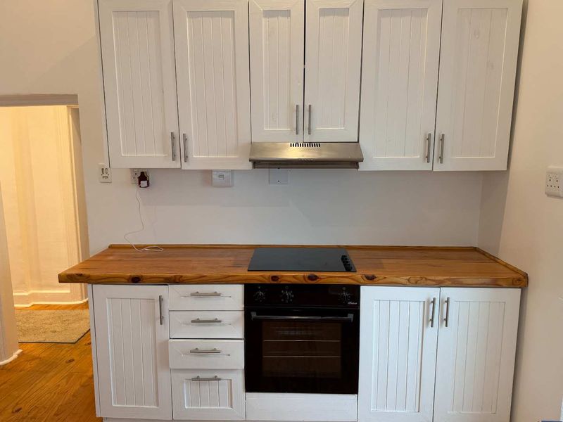A compact kitchen featuring white cabinetry, a natural wood countertop, and integrated appliances including an oven, hob, and extractor fan.