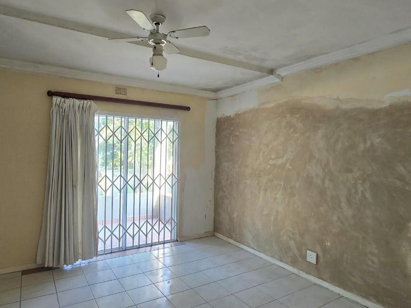 An empty living room featuring tiled flooring, a ceiling fan, and a sliding glass door with a security gate, requiring wall repairs and cosmetic updates.