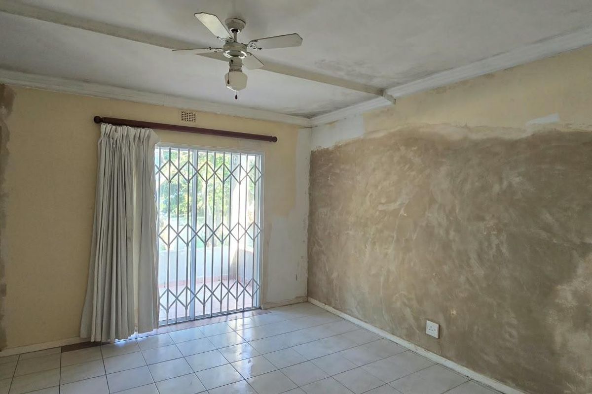 An empty living room featuring tiled flooring, a ceiling fan, and a sliding glass door with a security gate, requiring wall repairs and cosmetic updates.
