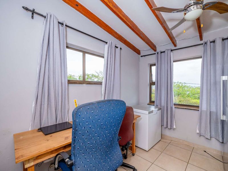 A functional office space featuring exposed timber ceiling beams, tiled flooring, and large windows that provide natural light and garden views.
