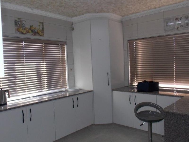 A functional kitchen featuring white built-in cabinetry, granite countertops, and tiled splashbacks, illuminated by natural light through large windows with venetian blinds.