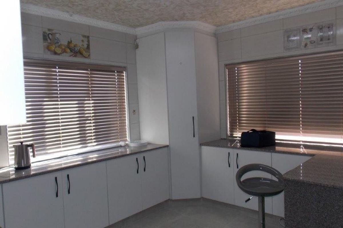 A functional kitchen featuring white built-in cabinetry, granite countertops, and tiled splashbacks, illuminated by natural light through large windows with venetian blinds.