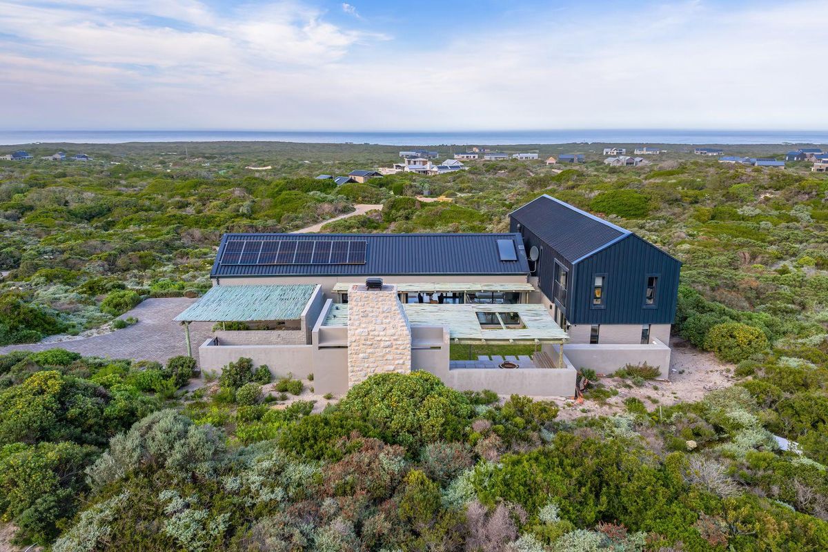 Aerial view of a modern coastal residence featuring a metal roof with solar panels, stone chimney, and integrated outdoor pergola area.