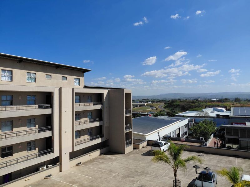 An elevated view of the apartment building exterior, featuring multiple balconies and an adjacent paved parking area with surrounding landscape views.