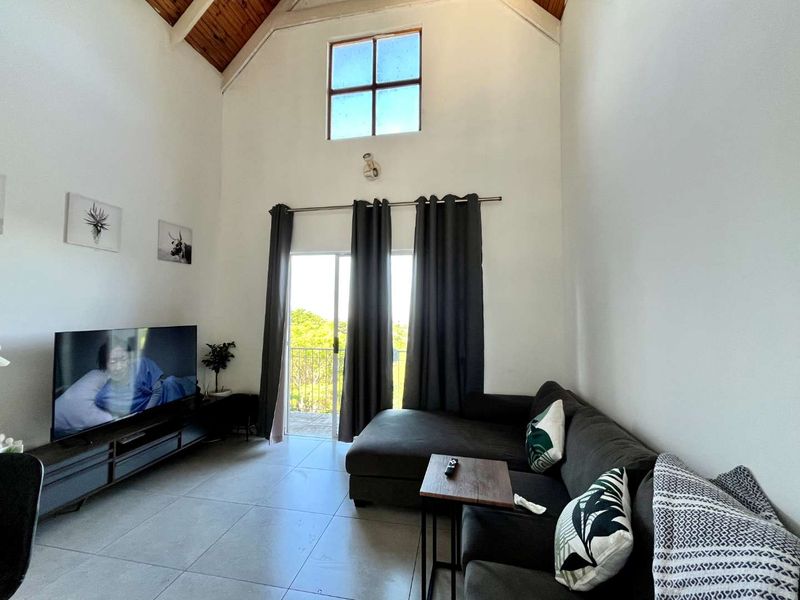 Spacious living room featuring a high vaulted timber ceiling, large sliding glass door leading to a balcony, and neutral tiled flooring.