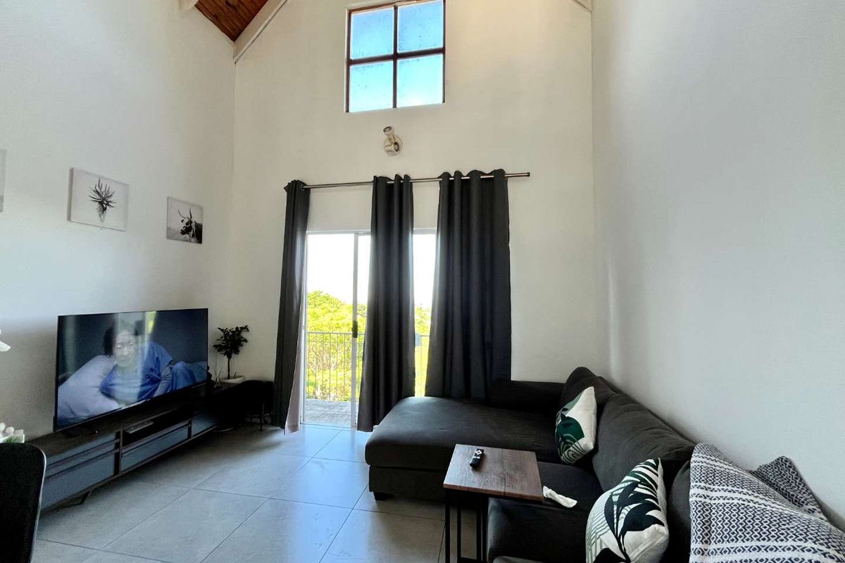 Spacious living room featuring a high vaulted timber ceiling, large sliding glass door leading to a balcony, and neutral tiled flooring.