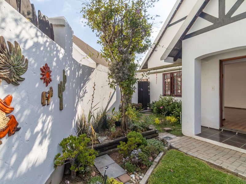 A well-maintained rear garden area featuring a paved walkway leading to the home entrance, bordered by a white boundary wall and established garden beds.