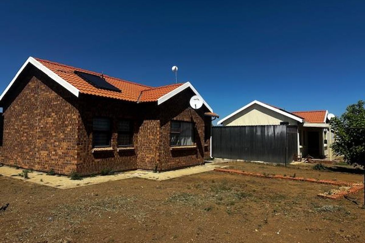 Exterior view of a face-brick residential property featuring a tiled roof, solar water heating panel, and an expansive, undeveloped garden area.