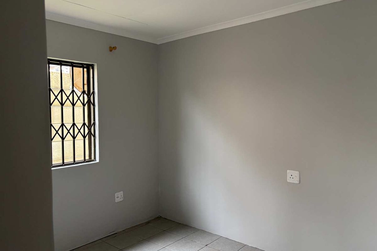 A clean, empty bedroom featuring neutral grey walls, tiled flooring, and a window fitted with a security gate.