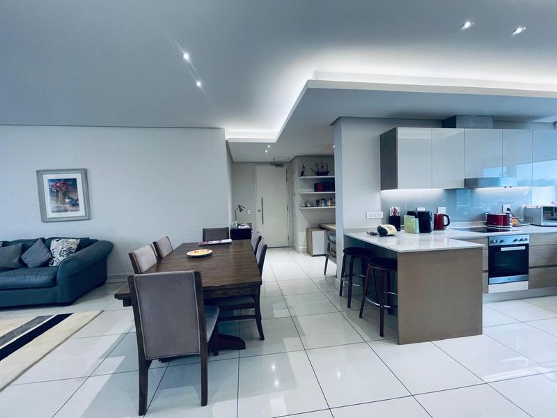 Modern open-plan living and dining area featuring a sleek kitchen with stone countertops, integrated appliances, and sophisticated recessed and cove lighting throughout.