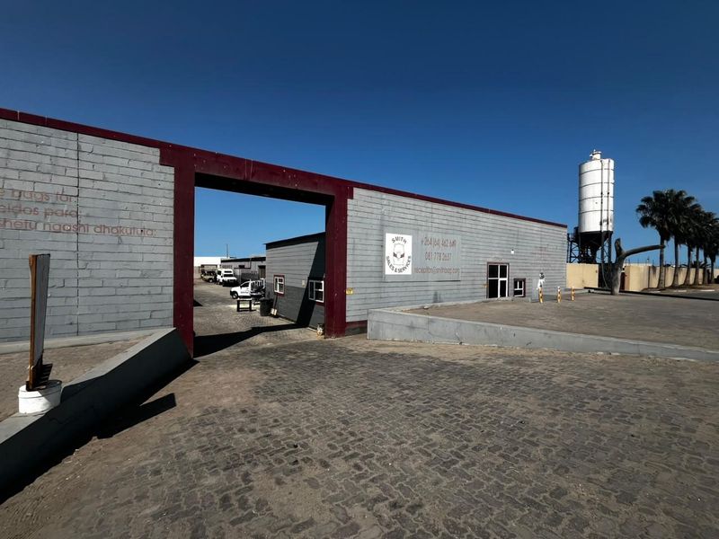 Industrial property entrance featuring a large steel gate frame, concrete block perimeter wall, cobblestone driveway, and an on-site silo.