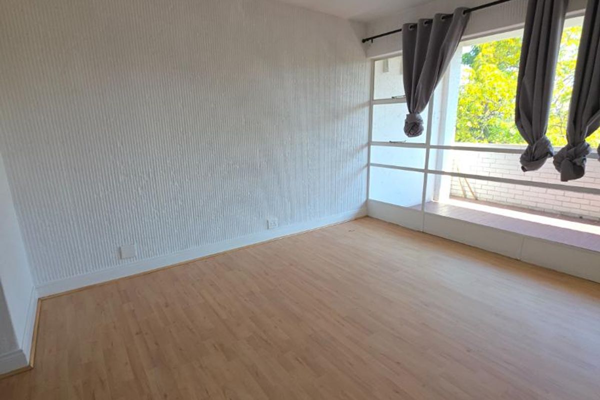 A bright, empty bedroom featuring light-toned laminate flooring, textured wall finishes, and a large window providing natural light.