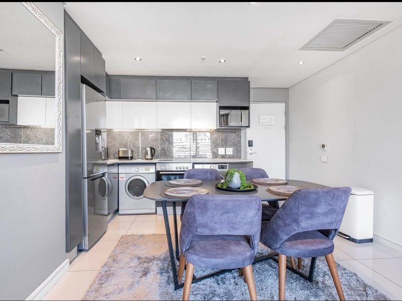 Modern open-plan kitchen and dining area featuring sleek built-in cabinetry, integrated appliances, tiled flooring, and recessed lighting in a contemporary apartment setting.