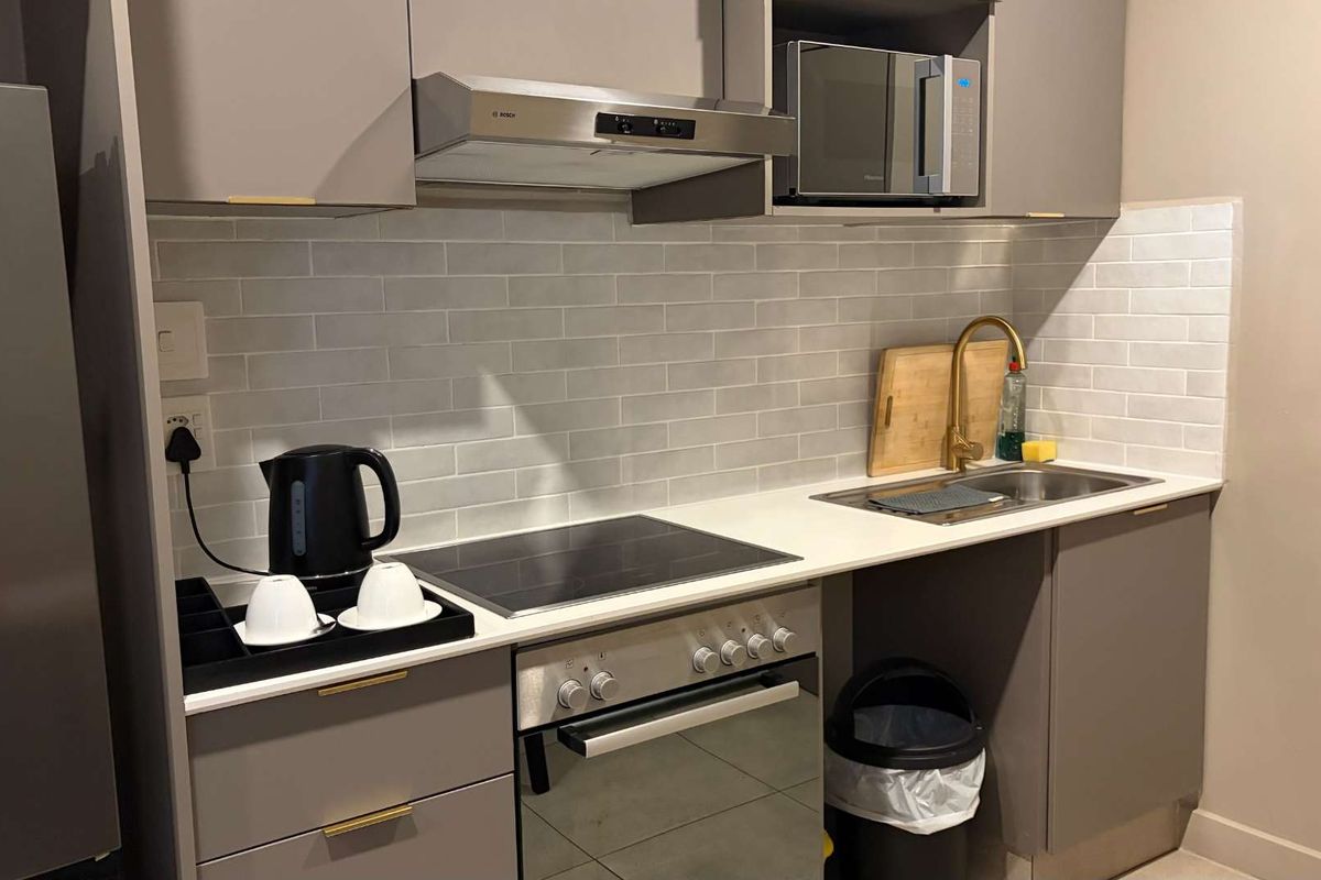 Modern compact kitchen featuring grey cabinetry, subway tile backsplash, integrated appliances including an oven, hob, extractor fan, and microwave with a brass faucet.