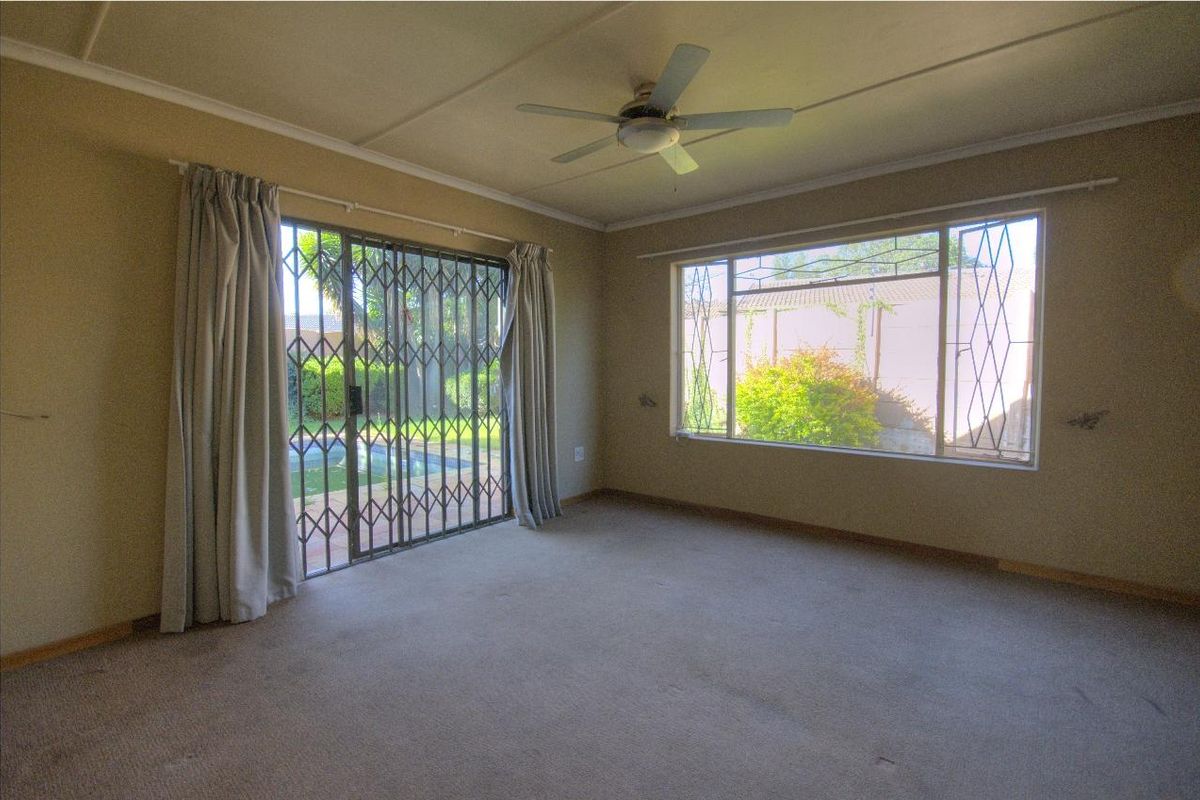 Spacious living room featuring neutral carpeting, a ceiling fan, and large windows with security gates providing access to the outdoor pool area.
