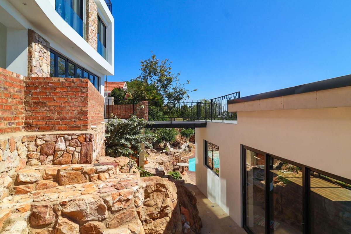 Exterior view of the property featuring stone retaining walls, a metal pedestrian bridge, and modern building architecture with large glass sliding doors.