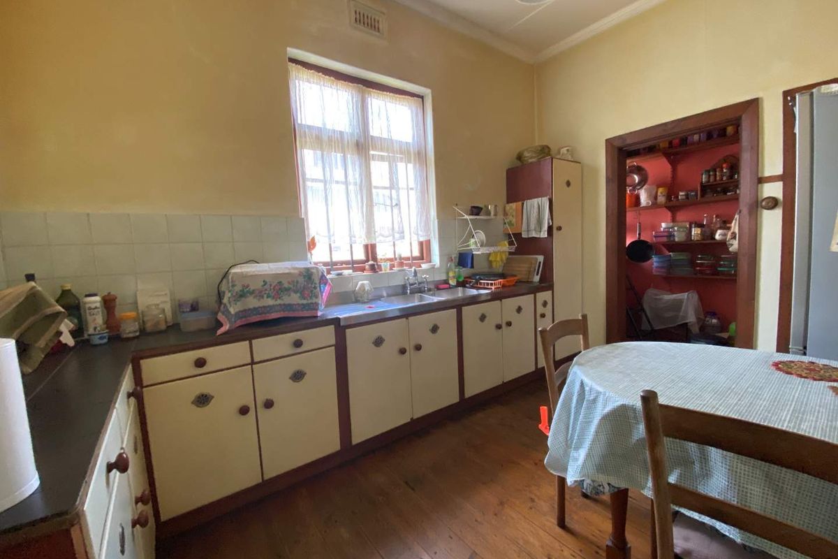 A dated kitchen featuring original timber cabinetry, a stainless steel sink, and exposed timber flooring, with an adjacent walk-in pantry area.