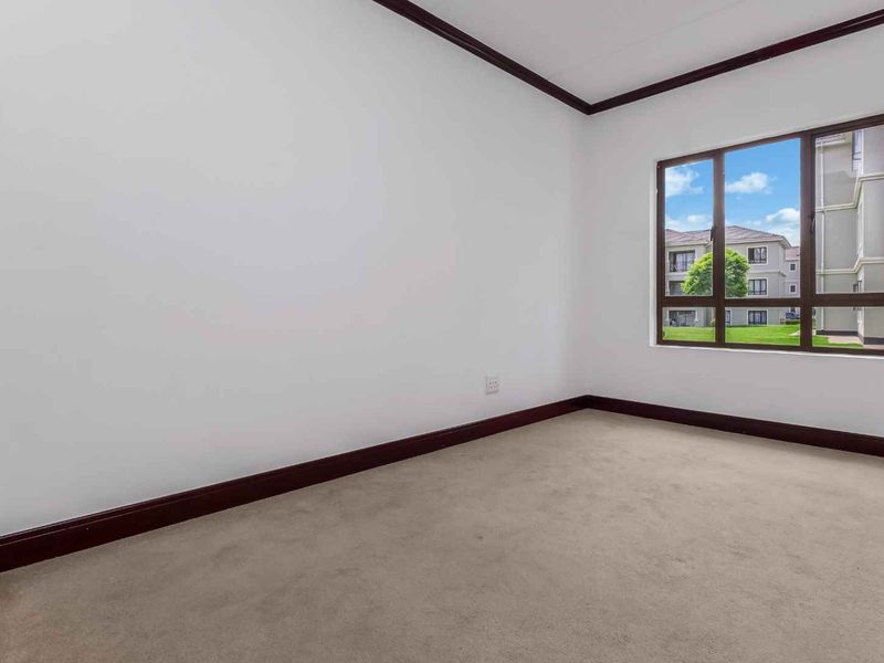 A bright, empty bedroom featuring neutral carpet flooring, dark wood trim, and a large window providing natural light and a view of the complex.