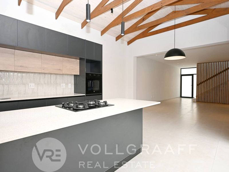Modern open-plan kitchen featuring a central island with gas hob, sleek cabinetry, and striking exposed timber trusses, flowing into a spacious living area.