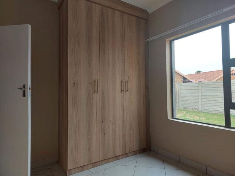 A bright, empty bedroom featuring modern wood-finish built-in wardrobes, neutral tiled flooring, and a large window overlooking the rear garden area.