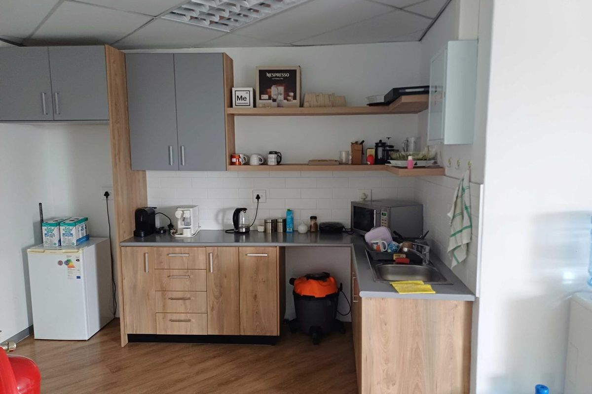 A functional office kitchenette featuring wood-grain cabinetry, grey upper cabinets, a stainless steel sink, and a tiled backsplash.