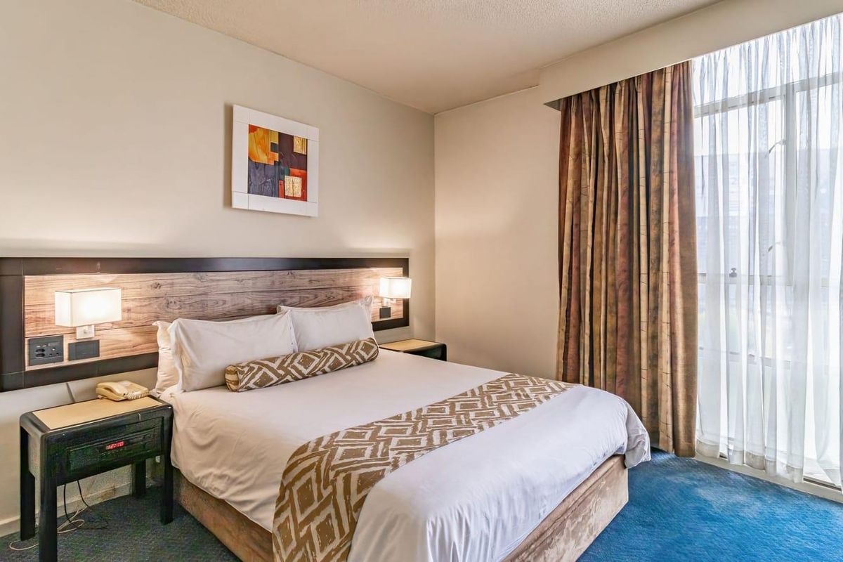 A comfortable bedroom featuring a built-in headboard with integrated lighting and large windows dressed with floor-to-ceiling curtains.