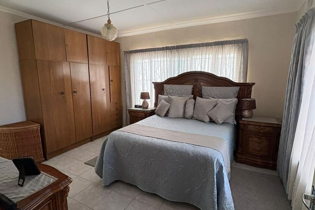A well-maintained bedroom featuring neutral tiled flooring, a large built-in wooden wardrobe, and a window fitted with security bars.