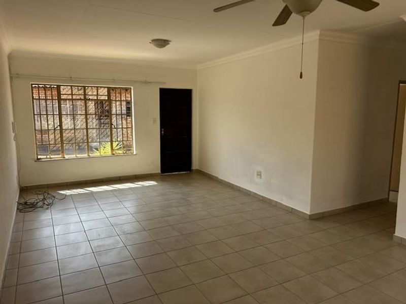 An empty, open-plan living area featuring neutral tiled flooring, a ceiling fan, and security bars on the window.
