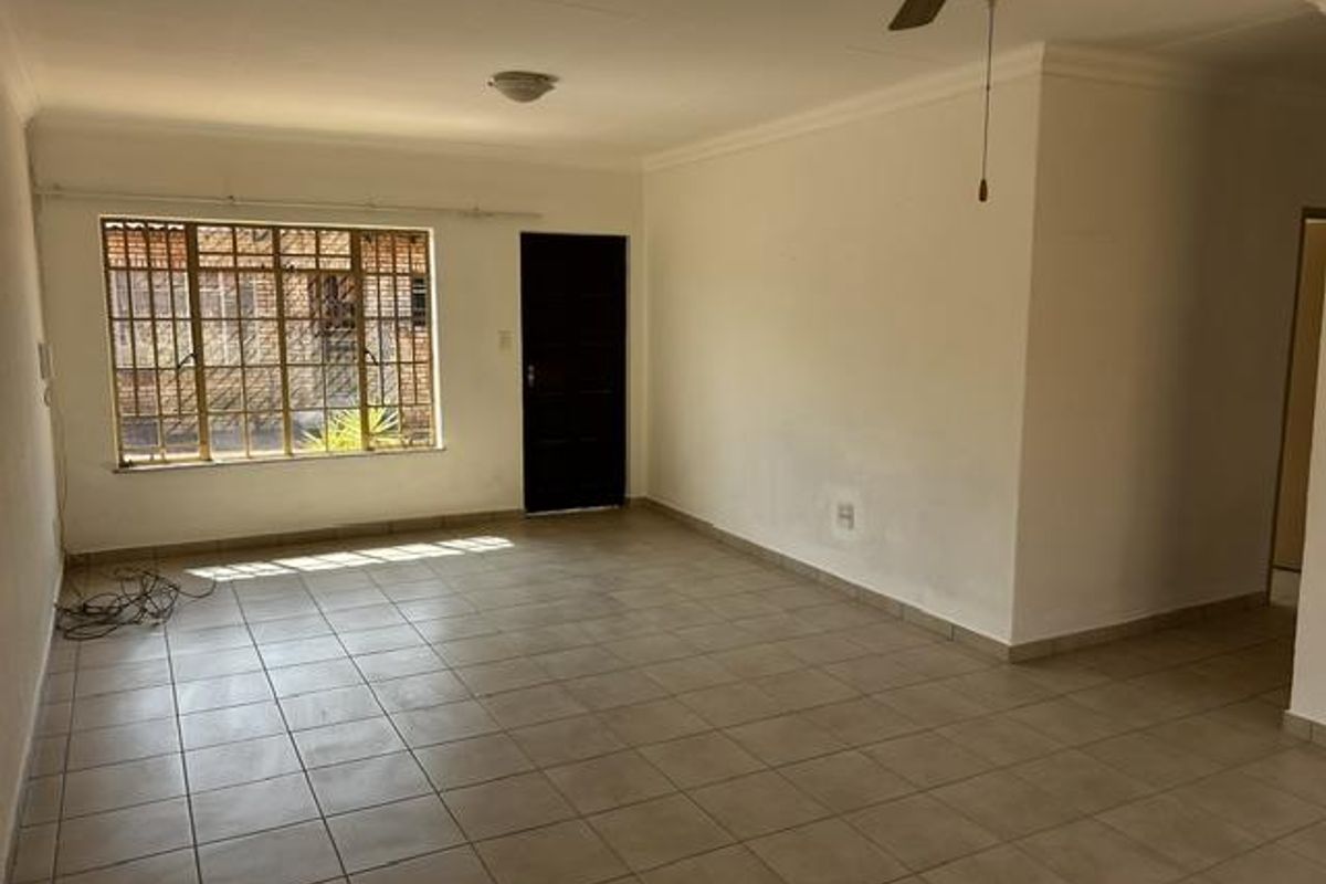 An empty, open-plan living area featuring neutral tiled flooring, a ceiling fan, and security bars on the window.