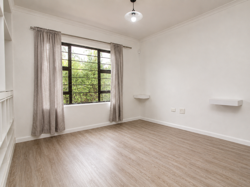 A bright, empty bedroom featuring light wood-look laminate flooring, built-in shelving, and a window with security bars.