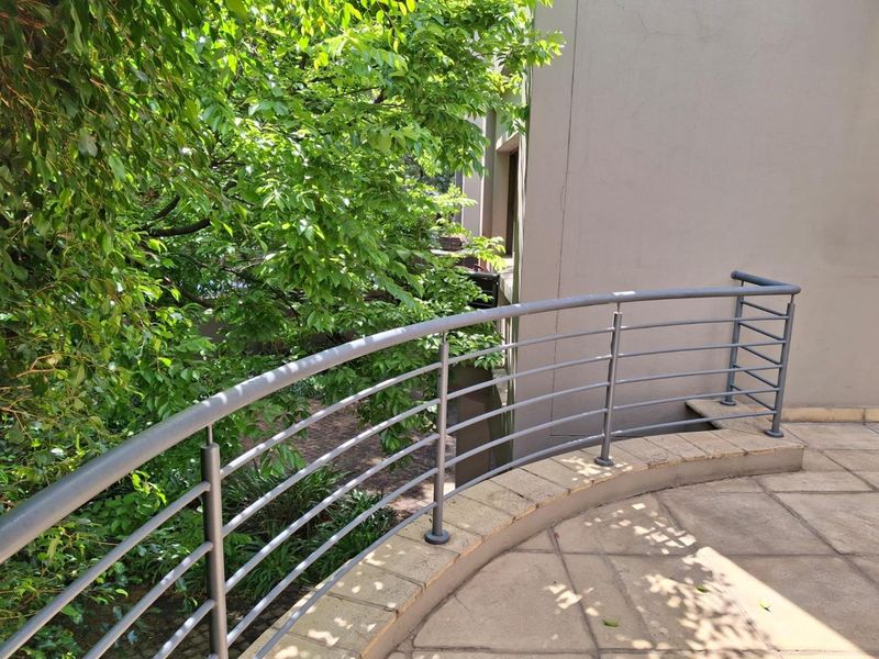 A curved balcony featuring a metal balustrade and paved flooring, overlooking lush greenery and the building exterior.
