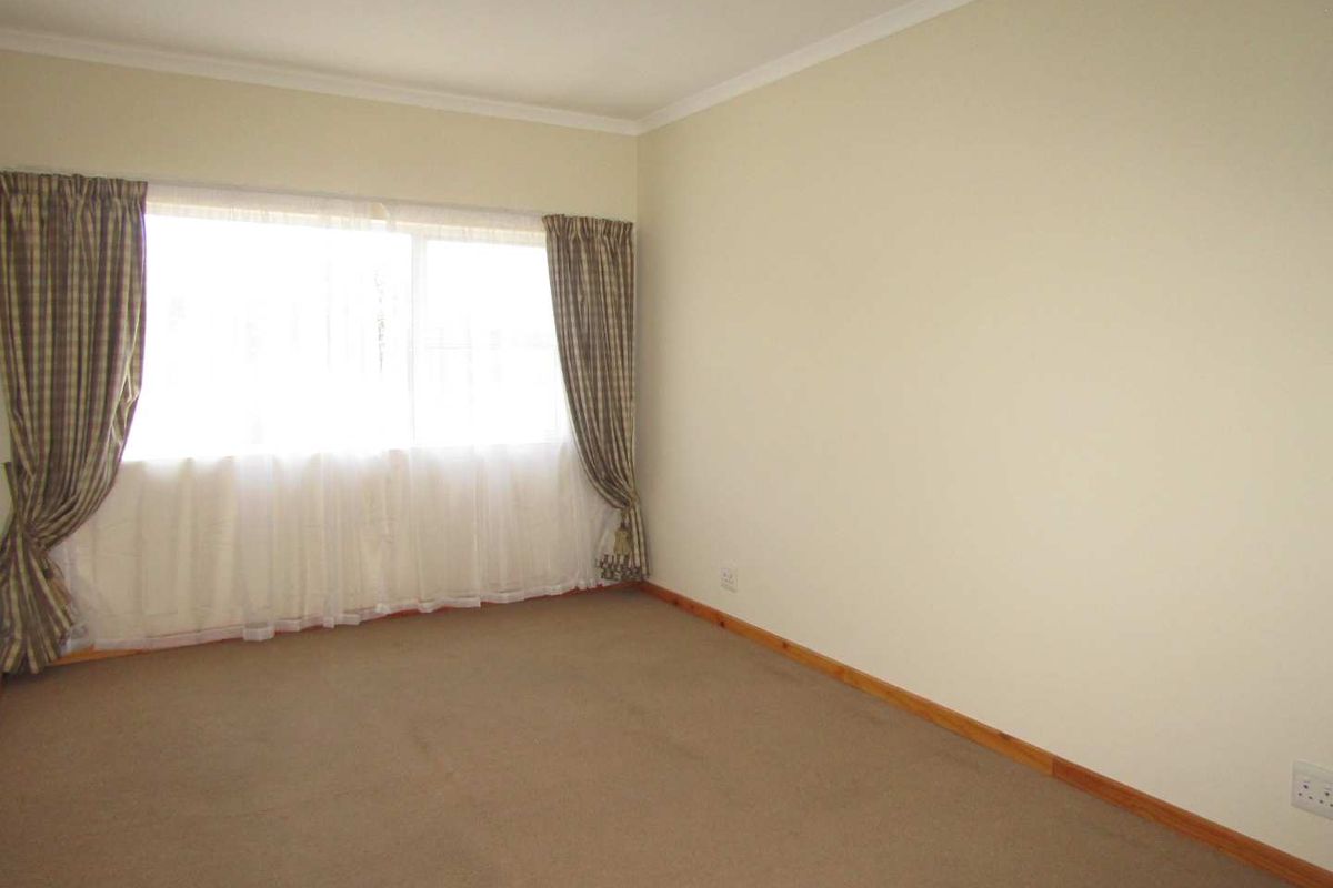 A spacious, empty bedroom featuring neutral wall tones, carpeted flooring, and a large window with natural light.